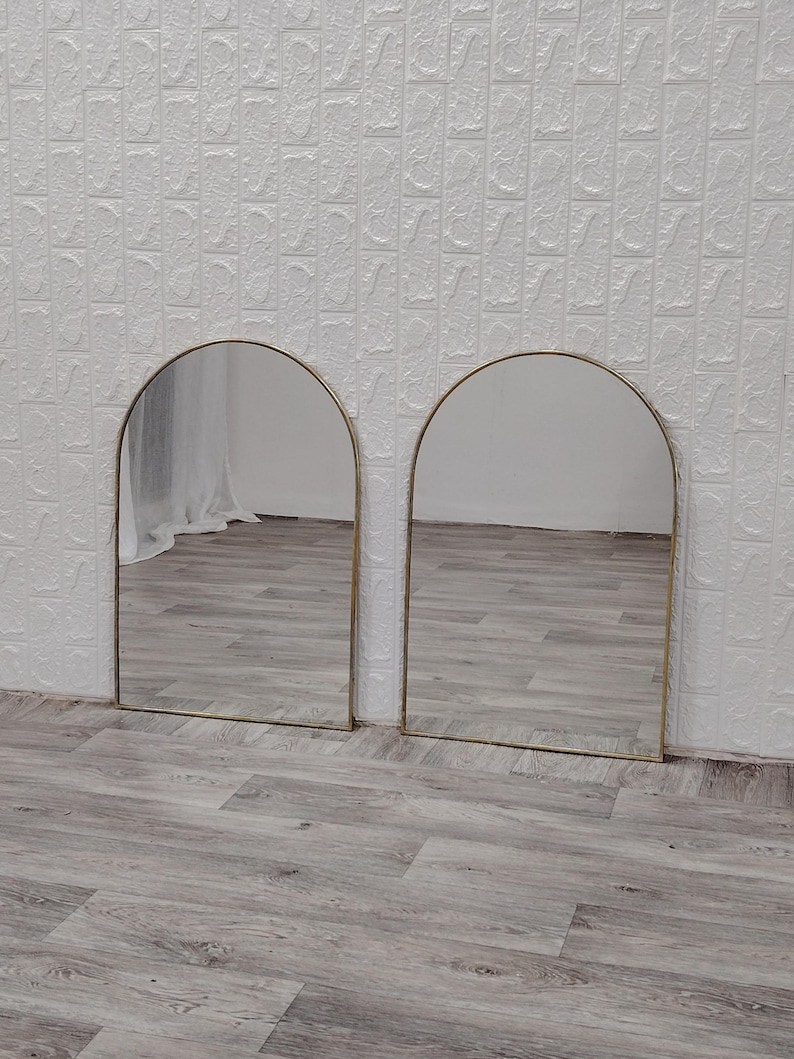 Op de afbeelding: Twee gouden boogspiegels met een witte achtergrond. De spiegels reflecteren de vloer, die van licht hout is.