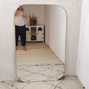 May include: A large, rectangular mirror with a rounded top and gold frame. The mirror is reflecting a person standing in front of it, as well as a white cabinet with a vase of dried flowers.