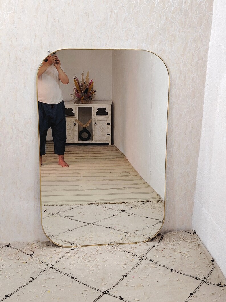 May include: A large, rectangular mirror with a rounded top and gold frame. The mirror is reflecting a person standing in front of it, as well as a white cabinet with a vase of dried flowers.