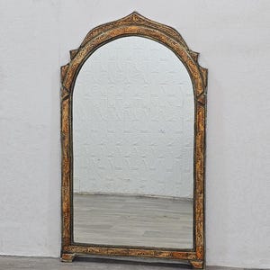 May include: A decorative arched mirror with a carved wooden frame. The frame is painted in a light brown color with intricate floral designs. The mirror is reflecting a white wall and a wooden floor.