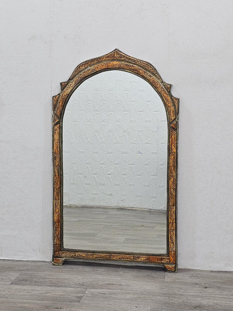 May include: A decorative arched mirror with a carved wooden frame. The frame is painted in a light brown color with intricate floral designs. The mirror is reflecting a white wall and a wooden floor.