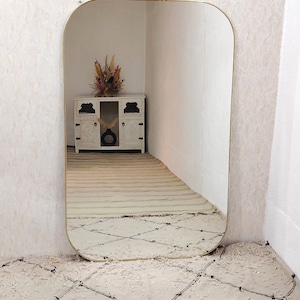 May include: A gold-framed, rounded rectangular mirror reflecting a white cabinet with a dried flower arrangement on top. The mirror is leaning against a wall with a white and black patterned rug in front of it.