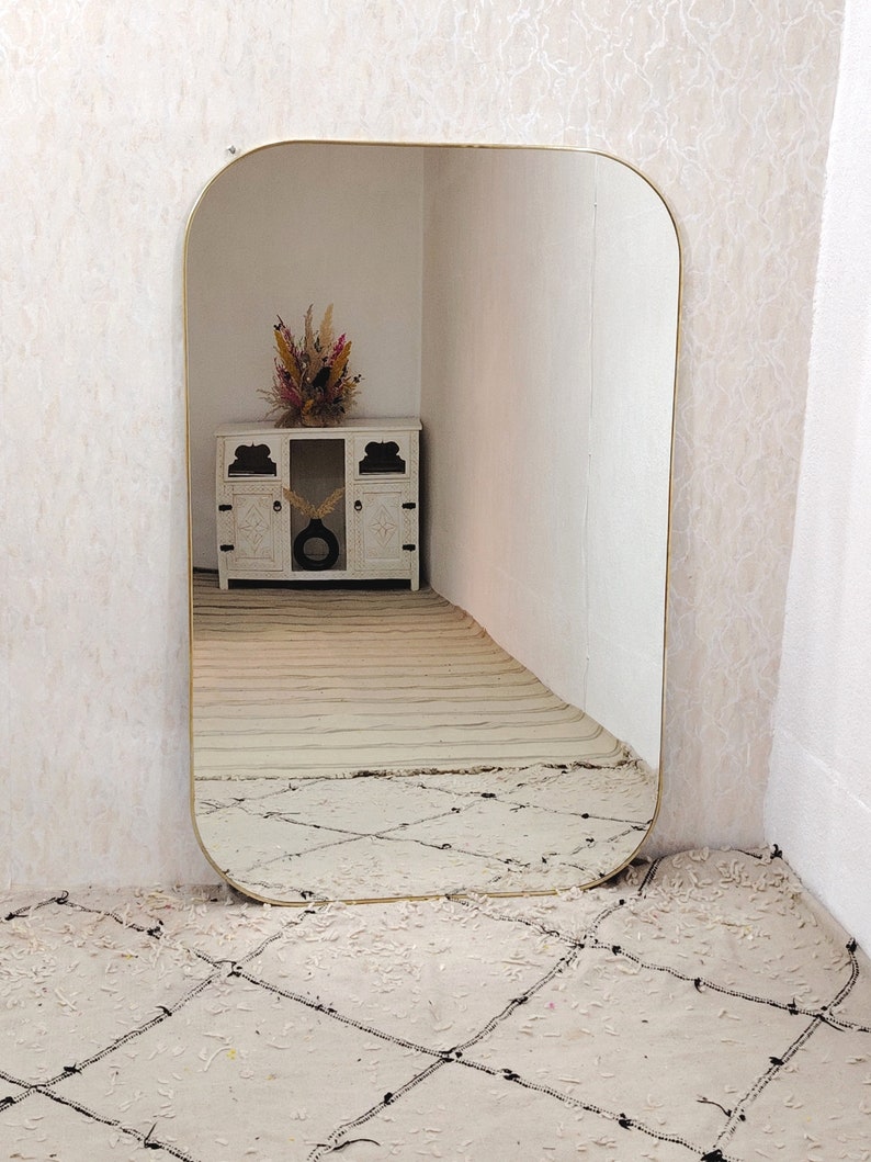 May include: A gold-framed, rounded rectangular mirror reflecting a white cabinet with a dried flower arrangement on top. The mirror is leaning against a wall with a white and black patterned rug in front of it.