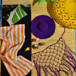 Könnte beinhalten: Ein Strand-Stillleben mit einer gelb-grünen aufblasbaren Ananas, einem lila Sonnenhut und einem gestrickten orange-weiß gestreiften Schal. Weitere Artikel sind ein grün-weißes Kissen, ein lila Netz und kleine dekorative Gegenstände.