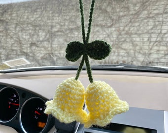 May include: A pair of yellow crocheted bells with green crocheted leaves hanging from a rearview mirror in a car.