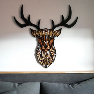 Op de afbeelding: Een geometrisch hertenkop wanddecor met zwarte geweien en een bruin en gouden patroonlichaam. De hertenkop is gemonteerd op een witte muur boven een grijs hoofdbord met twee kussens.