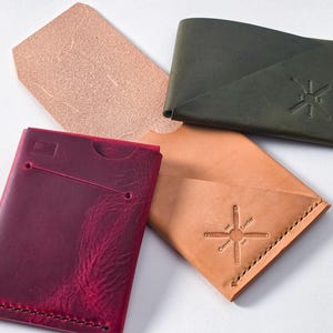 May include: A selection of leather wallets in shades of burgundy, olive green, and tan. Each wallet has a distinct design, with some featuring a compass emblem and detailed stitching. A separate piece of raw leather is also visible.