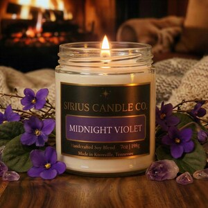 Pode incluir: Uma vela acesa num frasco de vidro, com o rótulo "Midnight Violet" da Sirius Candle Co. A vela é rodeada por flores e cristais roxos. O rótulo também diz "Handcrafted Soy Blend 198g."