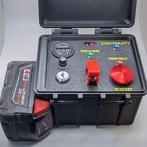 May include: A black, rectangular device with a hinged lid, featuring a digital voltage display, power and continuity indicators, and a large red button labeled "ARMED." A red and black battery pack is attached to the side.