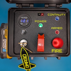May include: A black box with a red switch, a red button labeled "FIRE", and a digital display showing "8.8.8" with the label "VOLTAGE" and "DC V". The box also has a green light labeled "POWER", a blue light labeled "CONTINUITY", and a red light labeled "ARMED".