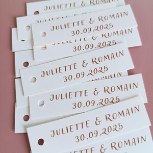 Peut inclure: Plusieurs étiquettes rectangulaires blanches empilées, imprimées avec les noms "JULIETTE & ROMAIN" et la date "30.09.2025" en encre marron. Chaque étiquette a un petit trou en haut. Le fond est rose uni.