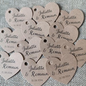 Peut inclure: Plusieurs étiquettes en forme de cœur en carton beige. Chaque étiquette est percée d'un petit trou et porte les noms "Juliette & Romain" et la date "31.08.2024" imprimés en noir. Les étiquettes sont disposées sur une surface texturée.