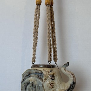May include: A ceramic elephant-shaped hanging planter featuring a cream and brown glazed finish. The planter is suspended by thick, braided beige cords adorned with two smooth, round wooden beads at the top, creating a functional decorative piece.