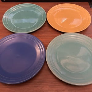 May include: A set of four round ceramic plates featuring a glossy finish and concentric ring detailing along the outer rim. The collection includes one blue plate, one yellow plate, and two light green plates, all arranged in a two-by-two grid.