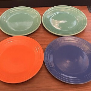 May include: A set of four round ceramic plates featuring a glossy finish. Two plates are light green, one is bright orange, and one is deep blue. Each plate has a series of concentric circular ridges embossed along the outer rim edge.