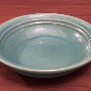 May include: A round, shallow ceramic bowl with a glossy, light turquoise glaze. The bowl features a smooth, curved interior basin and a distinct, raised circular ridge along the inner rim, creating a simple, functional design with a uniform finish.