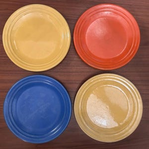 May include: A set of four round ceramic plates featuring a glossy finish and concentric ring detailing along the rims. The collection includes one bright yellow plate, one vibrant orange plate, one deep blue plate, and one muted mustard yellow plate.