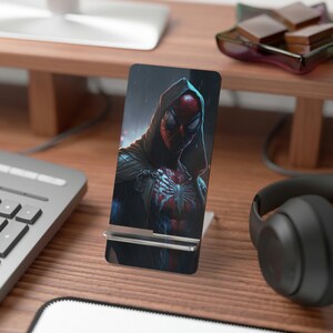 May include: A black phone stand with a graphic of a hooded Spiderman character with a red and black suit. The background is a dark blue with rain drops.