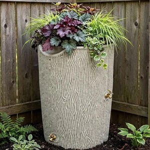 May include: A cylindrical rain barrel with a textured light gray exterior resembling tree bark. The barrel features two brass-colored spigots, one near the base and one higher up, and is filled with a variety of green and purple foliage.