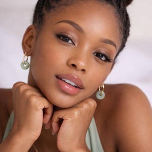 Peut inclure: Une femme à la peau foncée et aux cheveux tressés en chignon porte des boucles d'oreilles dorées avec des pendentifs circulaires vert jade. Elle porte un haut vert clair et un délicat collier en or. Ses mains sont sous son menton.