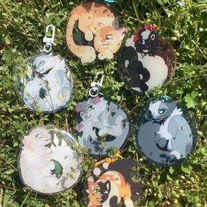 Op de afbeelding: Zeven ronde, transparante acryl sleutelhangers met kleurrijke cartoon kat illustraties. Elke sleutelhanger heeft twee katten in een knuffelpose. De katten zijn in verschillende kleuren, waaronder wit, grijs, zwart, oranje en bruin.