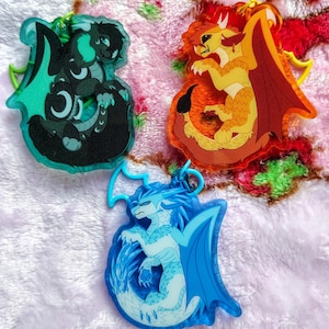 Puede incluir: Tres llaveros con temática de dragones. Uno es verde azulado y negro, otro naranja y amarillo, y el tercero azul y blanco. Cada llavero presenta un diseño de dragón con detalles intrincados y un lazo de color a juego.