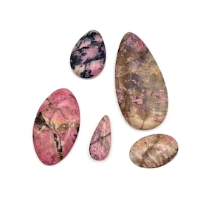 Op de afbeelding: Vijf gepolijste rhodoniet edelstenen in verschillende vormen. De stenen vertonen een mix van roze, zwarte en bruine tinten met unieke aderingpatronen. De edelstenen zijn geplaatst tegen een witte achtergrond.