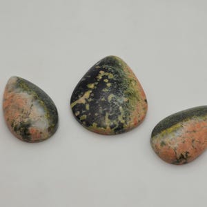 Puede incluir: Tres piedras de unakita pulidas con forma de lágrima. Las piedras muestran una mezcla de colores, incluyendo rosa, verde y negro. Las piedras están dispuestas sobre una superficie blanca.
