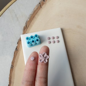 May include: A close-up of a hand holding a light pink flower-shaped polymer clay charm. The charm is on a white tile surface with a blue polymer clay mold and several light pink polymer clay circles.