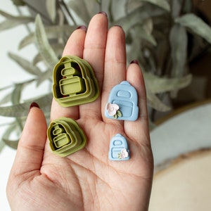 Puede incluir: Cuatro cortadores de galletas de diferentes tamaños en forma de mochila. Dos son verdes y dos son azules. Los cortadores azules tienen una pequeña flor rosa en la parte delantera.