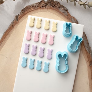 May include: A white surface with rows of pastel-colored bunny-shaped fondant decorations. There are four rows of bunnies in yellow, pink, purple, and blue. On the right side of the image are two blue plastic bunny-shaped cookie cutters.
