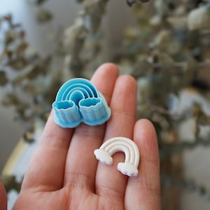 May include: Two rainbow-shaped cookie cutters, one blue and one white. The blue cutter has a more detailed design with multiple layers. The white cutter has a simpler design with clouds on either side.