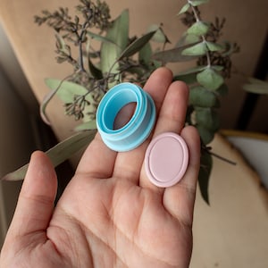 May include: A light blue plastic ring and a light pink oval-shaped plastic piece. The items are held in a person's hand.