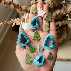 May include: A hand holding a variety of blue and green Christmas tree cookie cutters. The cutters are in different sizes and shapes.