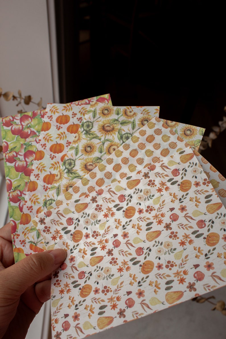 May include: Four sheets of decorative paper with autumnal themes. The papers feature different patterns including pumpkins, sunflowers, pears, apples, and leaves in shades of orange, yellow, brown, and red.