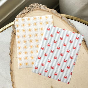 May include: Two patterned paper sheets. One features a repeating pattern of orange suns on a white background. The other has a pattern of red crabs on a blue and white striped background. The sheets are displayed on a wooden surface.