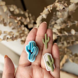 Puede incluir: Tres cortadores de galletas con forma de cactus, uno azul, uno blanco con un cactus verde y uno verde. Los cortadores están en una mano.