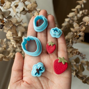 May include: A set of five cookie cutters in the shape of strawberries and flowers. The cutters are light blue and the strawberries are red with green leaves.