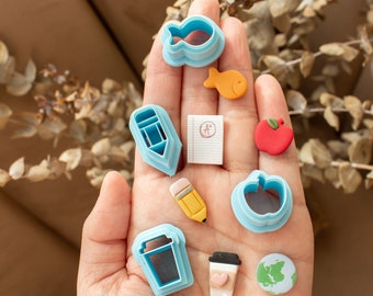 May include: A collection of blue plastic cookie cutters in the shape of a fish, pencil, coffee cup, apple, and a globe. There is also a small yellow pencil, a red apple, a small piece of paper with the letter "A" written on it, and a small orange fish.