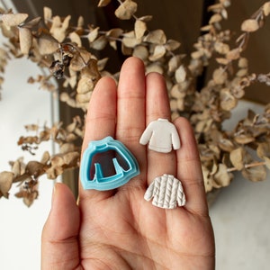 May include: Three sweater-shaped cookie cutters, one blue, one white, and one white with a textured design.