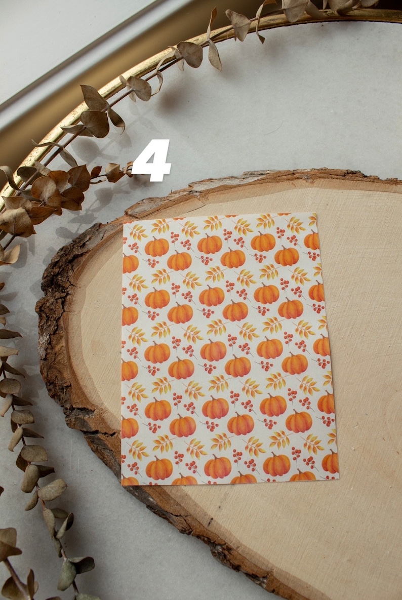 May include: A sheet of white paper with a repeating pattern of orange pumpkins, red berries, and yellow leaves. The paper is on a wooden surface.