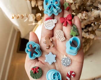 May include: A hand holding a variety of blue and white Christmas cookie cutters, including a gingerbread man, a snowflake, a candy cane, a mitten, a bell, a bow, a wreath, and a Christmas tree. There are also several small, colorful clay ornaments, including a Santa Claus, a red bow, a green holly sprig, and a candy cane.