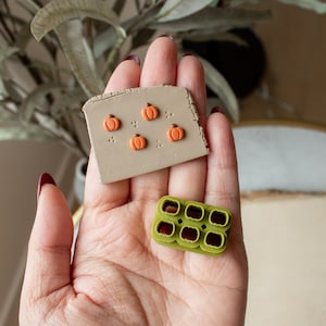 Puede incluir: Una mano sosteniendo dos herramientas de arcilla polimérica. Una es una lámina de arcilla beige con cinco formas de calabaza naranja. La otra es un molde de arcilla polimérica verde con ocho cavidades en forma de calabaza.