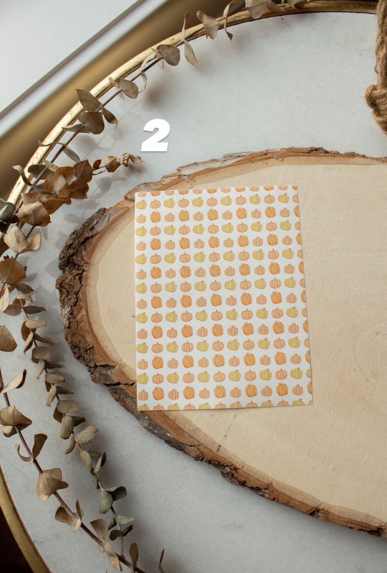 May include: A white card with a pattern of orange and yellow pumpkins on a wooden surface. The number "2" is written on the surface.