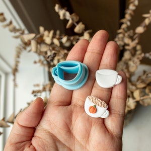 May include: A set of three cookie cutters in the shape of coffee mugs. The cutters are made of blue plastic and white fondant. One mug has a pumpkin spice design.