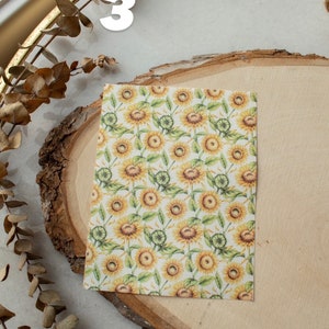 May include: A sheet of white paper with a repeating pattern of yellow sunflowers and green leaves. The paper is on a wooden slice.