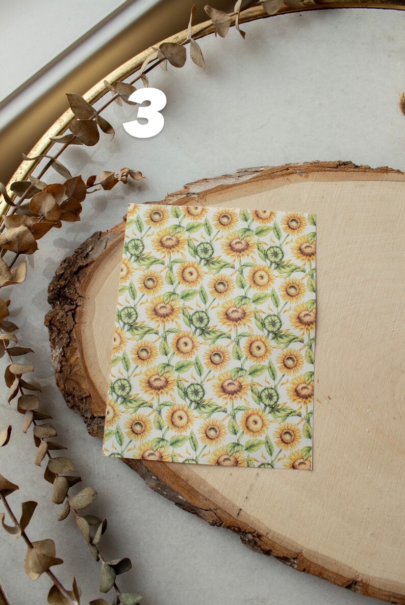 May include: A sheet of white paper with a repeating pattern of yellow sunflowers and green leaves. The paper is on a wooden slice.