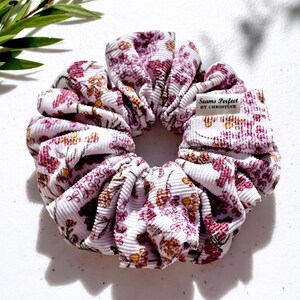 以下が含まれることがあります： ピンクと黄色の花柄の白いシュシュ。シュシュはコーデュロイ生地でできており、小さなタグには「Seams Perfect by Christine」と書いてあります。