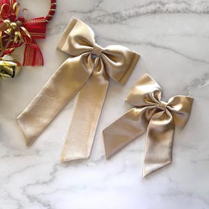 May include: Two gold satin hair bows with long tails. The larger bow is on the left and the smaller bow is on the right.