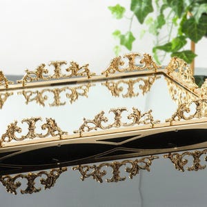 Könnte beinhalten: Ein goldfarbenes, verspiegeltes Tablett mit einem verzierten, dekorativen Rand. Das Tablett ist rechteckig und hat eine reflektierende Oberfläche.